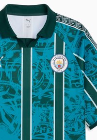 Φανέλα ποδοσφαίρου της Manchester City με κάθετες ρίγες σε τιρκουάζ και σκούρο πράσινο, αφηρημένα σχέδια, λογότυπο Puma και έμβλημα του συλλόγου στο στήθος.