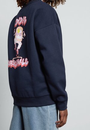 Personne portant un sweat-shirt bleu marine avec un personnage d'œuf de dessin animé coloré en train de faire du skateboard, avec un texte rose « ADIDAS ORIGINALS » dans le dos.