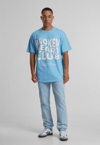 Camiseta oversize de color azul claro con un atrevido texto blanco "BROKEN HEART CLUB" acompañado de gráficos de nubes, combinada con unos jeans de denim claros y zapatillas.
