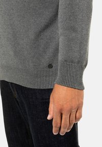 Grauer Strickpullover mit gerippten Bündchen und einem runden schwarzen Logodetail. Vorgestellt mit dunklen Jeans und einer Nahaufnahme des strukturierten Stoffes.
