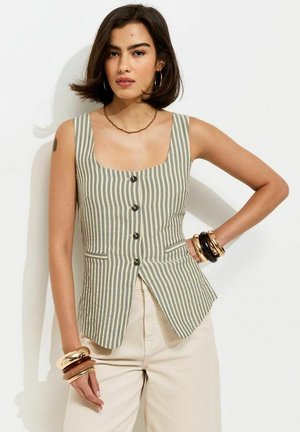 Vrouw met kort donker haar draagt een mouwloze top met strepen en knopen en lichte broek, geaccentueerd met oorringen en meerdere armbanden.