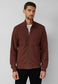 Gestrickte Reißverschlussjacke in dunklem Burgunderrot mit strukturiertem Muster, hohem Kragen und gerippten Bündchen. Dazu ein weißes T-Shirt darunter kombiniert.