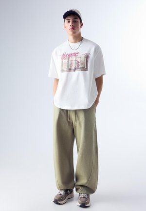 Jeune homme portant un t-shirt blanc à motifs, un pantalon ample vert olive, des baskets grises et une casquette blanche, debout les mains derrière le dos.