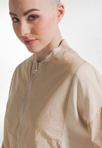 Giacca bomber beige con chiusura a zip, caratterizzata da una finitura texturizzata e un colletto a listino. Tessuto liscio e maniche a contrasto.