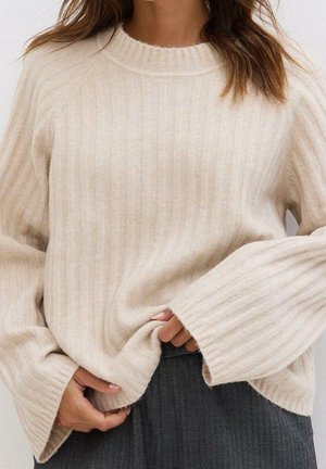Femme portant un pull en maille côtelée beige avec des manches oversize et un pantalon gris en chevrons, ajustant l'ourlet du pull.