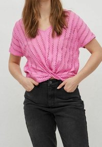 Růžový pletený top s výstřihem do V, zdobený vertikálními vykrojenými vzory, uvázaný v pase; kombinovaný s černými vysoce pasovými džíny.