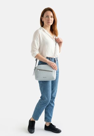 Woman with red hair wearing a white blouse, blue jeans, black sneakers, and holding a light gray crossbody bag.