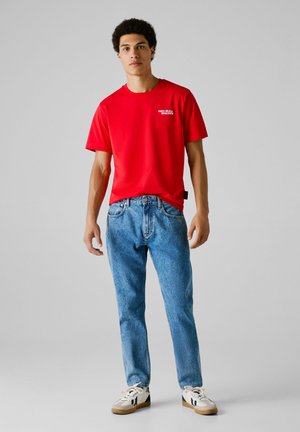 Ung mann iført en rød "Red Bull Racing" t-skjorte, blå jeans og hvite sneakers, står foran en lys grå bakgrunn.
