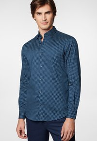 Granatowa koszula zapinana na guziki, wykonana z fakturowanej tkaniny, z małym białym wzorem w kropki, z ostrym kołnierzykiem i długimi rękawami.