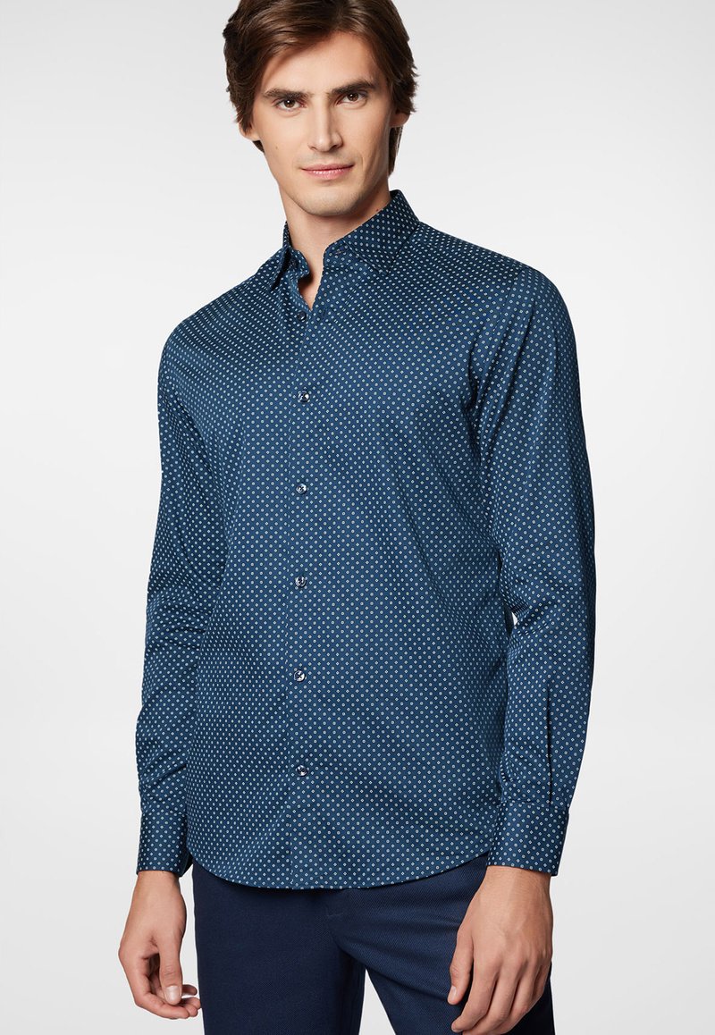 Granatowa koszula zapinana na guziki, wykonana z fakturowanej tkaniny, z małym białym wzorem w kropki, z ostrym kołnierzykiem i długimi rękawami.