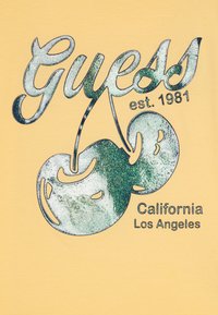 Dzeltena T-kreklā ar mirdzošo ķiršu grafiku un tekstu "Guess est. 1981 California Los Angeles" uzrakstītā treknajā melnajā fontā.