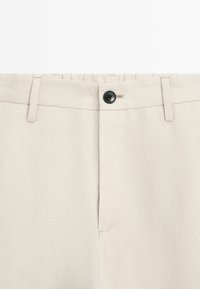Pantalon beige avec une fermeture par bouton noir, des passants pour ceinture et un design avant épuré avec des détails de couture subtils.