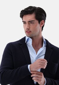 Mann, der den Bund eines dunkelblauen strukturierten Blazers über einem hellblauen Hemd anpasst, während er zur Seite blickt, vor einem schlichten weißen Hintergrund.