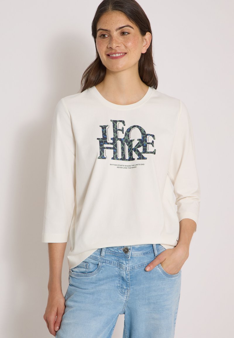Femme portant une chemise blanche à manches longues avec le texte "I GO HIKE" et un jean bleu clair, souriante, une main dans sa poche.