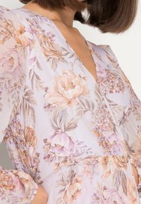 Ljust lila blommig klänning med puffärmar. Har en V-ringning och en åtsittande midja. Tyget har en genomskinlig, mjuk textur med pastellfärgade blommönster.