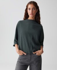 Vrouw met lang bruin haar, gekleed in een losse, donkere, gestructureerde trui en grijze jeans, staande met haar handen in haar zakken tegen een effen achtergrond.