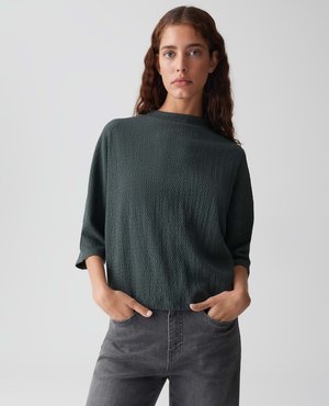 Eine Frau mit langen braunen Haaren, die einen dunklen strukturierten lockeren Pullover und graue Jeans trägt, steht mit den Händen in den Taschen vor einem einfarbigen Hintergrund.