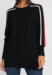 Pull en maille noire avec une texture côtelée, présentant des rayures rouges et blanches sur les épaules. Manches longues et ourlet droit.