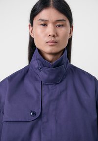 Lilla jakke med høy krage, knappeåpning og en jevn tekstur. Utformet med en enkel brystlomme og minimalistisk stil.