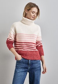 Maglione lavorato a maglia con collo alto in gradazione di crema e rosa, con una parte inferiore texturizzata rossa e polsini a costine. Abbinato a dei jeans.