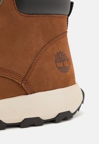 Bruine suède laars met zwarte accenten op de bovenkant, een witte rubberen zool en een gestempeld boomlogo op de zijkant.