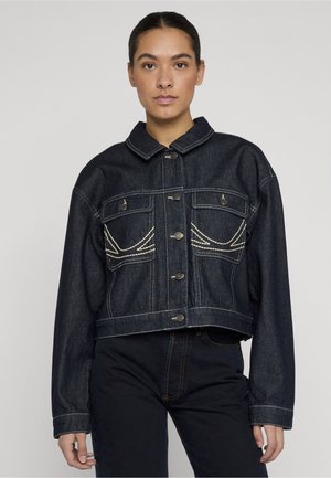 Mujer con cabello oscuro, lleva una chaqueta corta de mezclilla oscura con patrones bordados en blanco y pantalones oscuros de talle alto, de pie frente a un fondo liso.