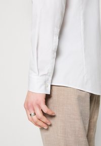 Main gauche avec une bague en argent sur le côté, portant une chemise blanche à manches longues et un pantalon beige texturé sur fond blanc.