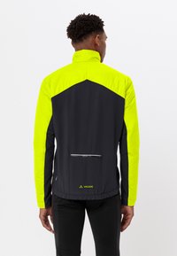 Uomo che indossa una giacca nera e gialla neon, in piedi con le spalle rivolte, mostrando una tasca con zip e il logo Vaude sulla parte bassa della schiena.