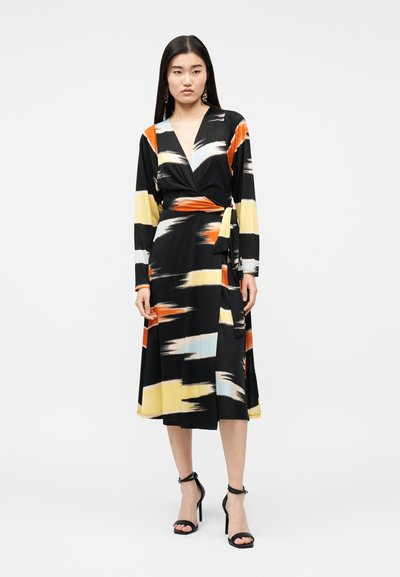 Γυναίκα που φοράει μαύρο φόρεμα τύπου wrap με αφηρημένα σχέδια σε πορτοκαλί, κίτρινο, λευκό και ανοιχτό γαλάζιο χρώμα, και μαύρα σανδάλια με ψηλό τακούνι.