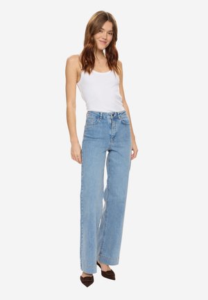 Tânără îmbrăcată într-un maiou alb, blugi largi și evazați, cu talie înaltă, de culoare albastru deschis, și pantofi cu toc înalt, cu vârf ascuțit, de culoare închisă, stând pe un fundal alb.