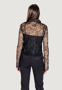 Top in pizzo nero con maniche lunghe, motivo floreale e schiena trasparente. Presenta uno strato di base nero opaco e un colletto. Pantaloni neri.