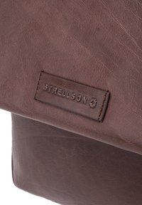 Bolso mensajero de cuero marrón con una superficie texturizada y forma rectangular. Incluye un parche con el logo cosido para identificación de la marca.