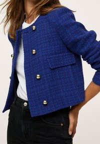 Veste en tweed bleu à coupe courte, boutons dorés et une poche poitrine unique, associée à un haut blanc et un pantalon foncé.