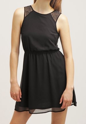 Zwarte mouwloze jurk met een strakke taille, doorschijnende mesh schouders versierd met studs, en een vloeiende rok die boven de knie valt.