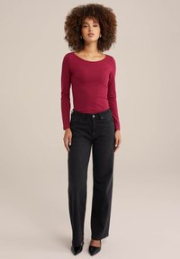 Langes burgunderrotes Oberteil mit einem U-Boot-Ausschnitt, kombiniert mit hochgeschnittenen schwarzen Jeans. Die Jeans haben einen klassischen Schnitt und dezente Hardware-Akzente.