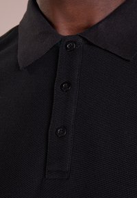 Zbliżenie osoby noszącej czarną, teksturowaną koszulkę polo z kołnierzykiem i trzema czarnymi guzikami zapiętymi przy szyi.