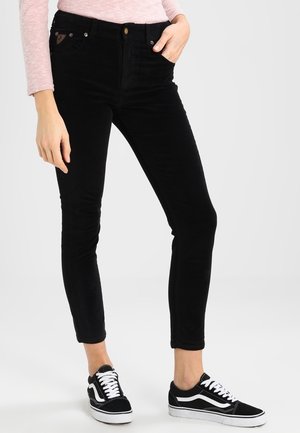 Vrouw die zwarte, nauwsluitende broek draagt, een lichtroze top met lange mouwen en zwart-witte sneakers, staand met één hand in haar zak.