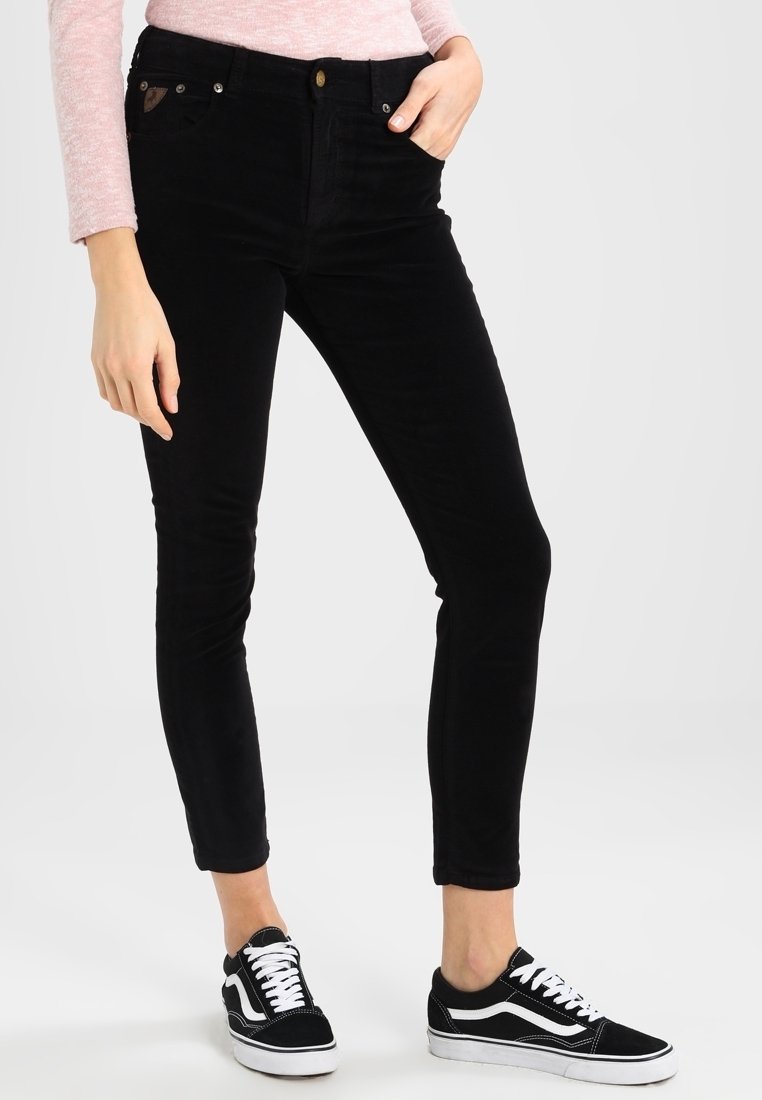 Femme portant un pantalon noir moulant, un haut à manches longues rose clair et des baskets noires et blanches, debout avec une main dans la poche.