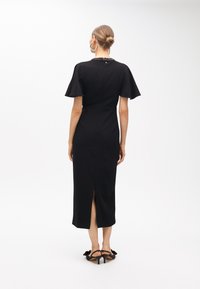 Kvinna med håret i en knut bär en svart midiklänning med korta fjärilsärmar, dekorerad halsringning, slits på baksidan och svarta låga klackar slingbacks.