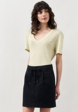 Γυναίκα με σγουρά σκούρα μαλλιά που φοράει ανοιχτό κίτρινο τοπ με V-λαιμόκοψη και κοντά μανίκια και μαύρη φούστα με κορδόνι, που στέκεται σε απλό φόντο.