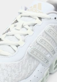 Hvit sportssko med mesh-overdel, forsterkede hull for lisser, tre teksturerte side-striper og en lys grønn mellomsåle. Har nøytrale lisser.