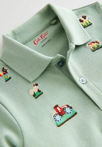 Polo vert clair avec des motifs brodés sur le thème de la ferme, y compris un tracteur et des animaux. Il possède un col et des boutons bleus.