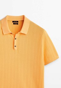 Polo à manches courtes de couleur orange vif, avec un motif en maille texturé, un plastron à deux boutons et un col classique.