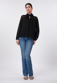 Blouse noire en tissu texturé, manches longues et encolure décolletée avec une ouverture, associée à un jean flare bleu et des sandales noires à brides.