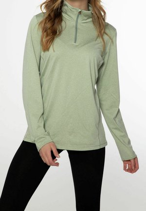 Kvinde iført en lysegrøn langærmet quarter-zip pullover og sorte leggings, stående mod en ensfarvet hvid baggrund.