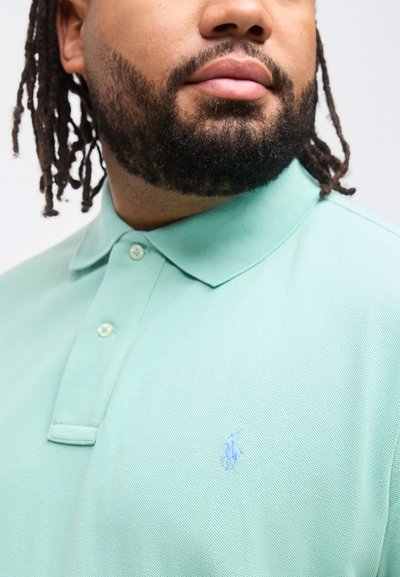 Ανδρας με μούσι και σκοτεινά μαλλιά που φοράει ανοιχτοπράσινο πόλο μπλουζάκι με κεντημένο μπλε λογότυπο αλόγου κ αι αναβάτη στο στήθος.