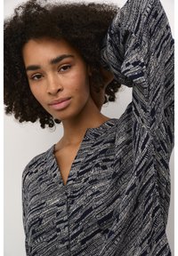 Blusa blu navy con motivo astratto bianco, maniche lunghe e scollatura a V. Il tessuto sembra morbido con una vestibilità larga e rilassata. Dettagli con bottoni visibili.