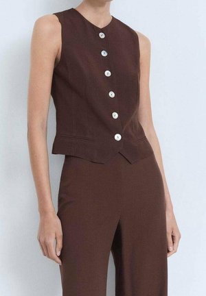 Ärmellose braune Weste mit sechs weißen Knöpfen, kombiniert mit passenden hoch taillierten braunen Hosen, getragen von einer stehenden Person vor hellem Hintergrund.