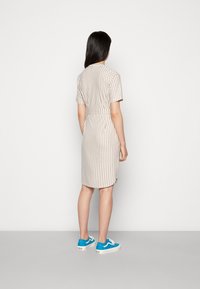 Pasiasta beżowa sukienka z krótkimi rękawami, z paskiem w talii, sięgająca do kolan, zestawiona z niebieskimi sneakersami. Prosty design widziany z tyłu.