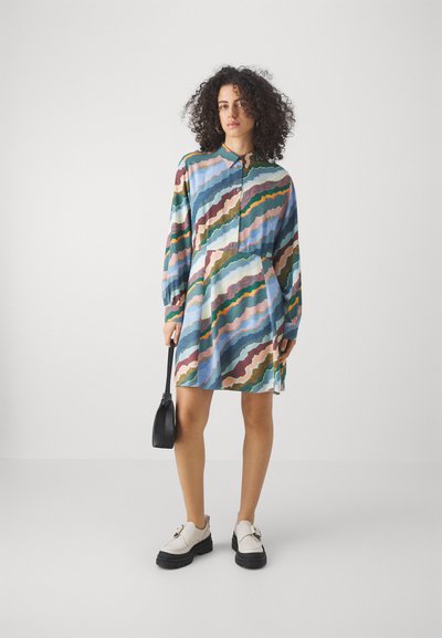 Abito a maniche lunghe in tessuto leggero con motivi a onde multicolori. Presenta una chiusura con bottoni davanti, colletto e una gonna svasata. Abbinato a una piccola borsa nera e scarpe bianche con suola spessa.